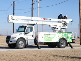 A white, electric bucket truck with the boom lowered and stowed, side stabilizers deployed parked below power lines