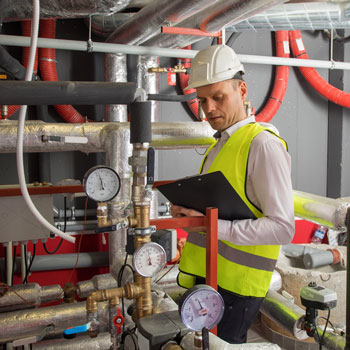 Man in hardhat and safety vest holding a clipboard inspecting a boilerroom with several pipes, hoses and guages visible throughout the photo