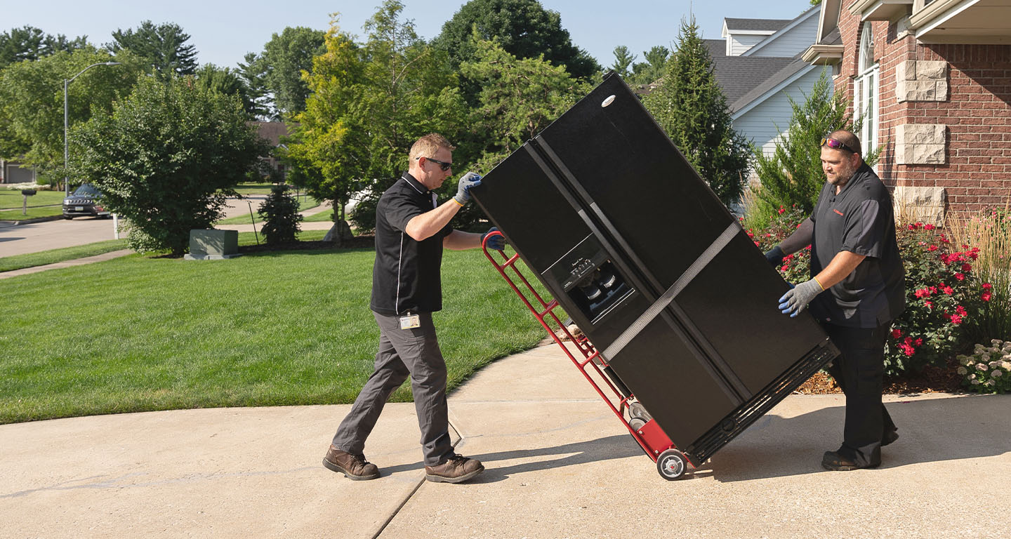 Two men carrying a refrigerator on a dolly in a residential driveway