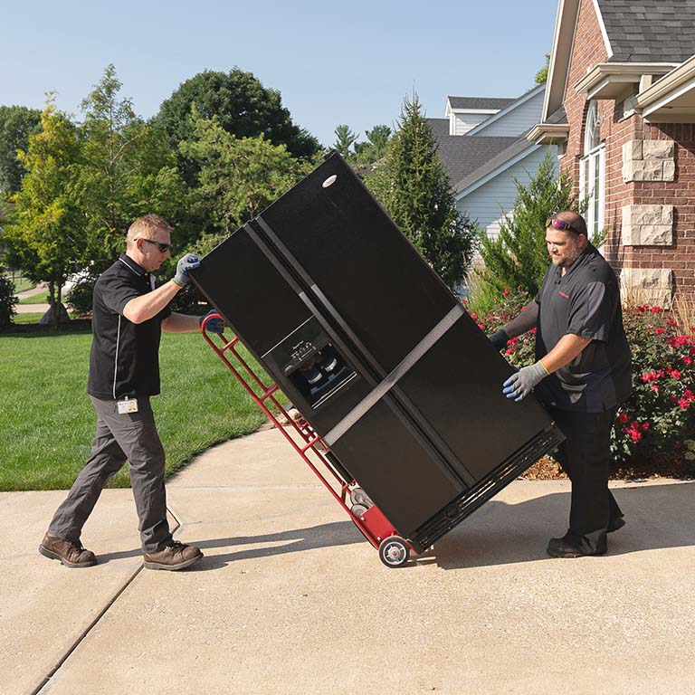 Two men carrying a refrigerator on a dolly in a residential driveway