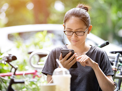 woman wearing glasses and a gray shirt sitting at a table outside smiling at her cell phone